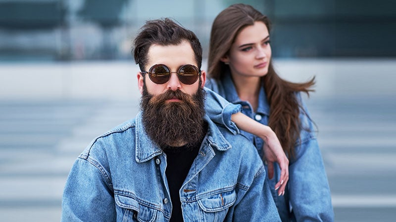 Close Up Portrait Of A Hipster Couple Of A Brutal Bearded Male In Sunglasses And His Girlfriend Dressed In Jeans Jackets Against Skyscraper.