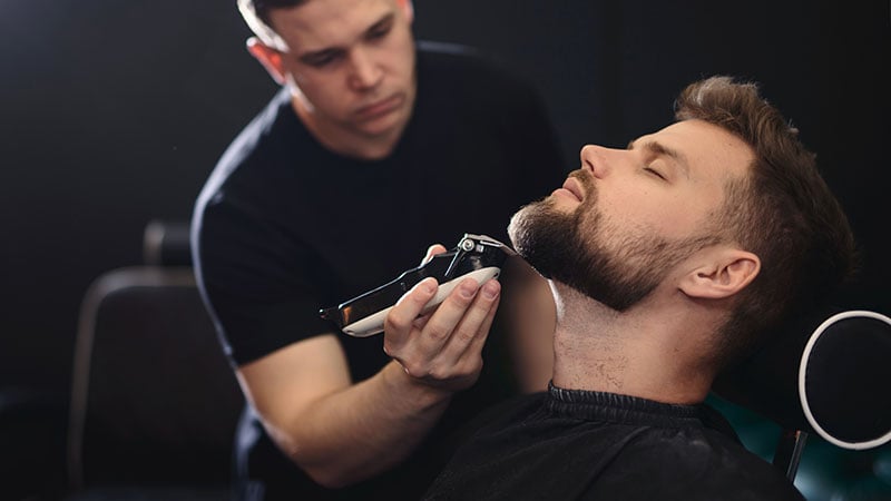 Getting Perfect Shape. Close Up Side View Of Young Bearded Man Getting Beard Haircut By Hairdresser Or Barber At Barbershop