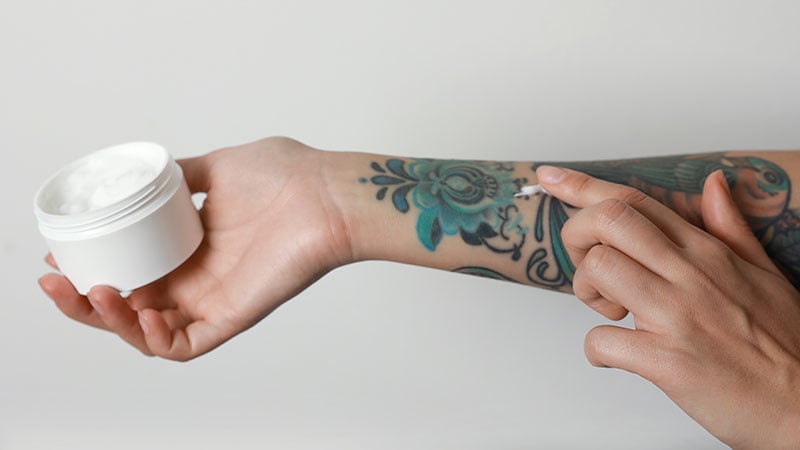 Woman Applying Cream On Her Arm With Tattoos Against Light Backg