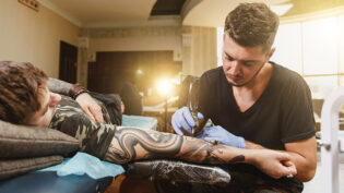 Professional Tattooer Artist Doing Picture On Hand Of Man By Machine Black Ink From A Jar. Tattoo Art On Body. Equipment For Making Tattoo Art. Master Makes Tattooed In Light Studio.