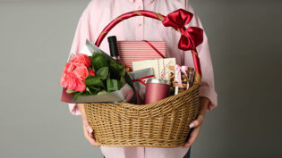 Woman Holding Wicker Basket Full Of Presents On Grey Background,