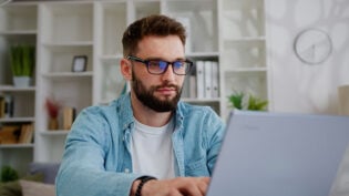 Handsome Caucasian Man Working On Laptop Computer While Sitting Behind Desk In Living Room. Freelancer Male Professional Writing An Important Email From Working From Home.